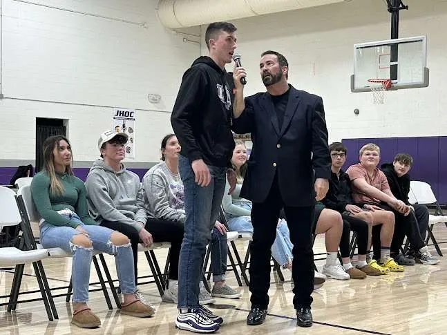 High school gym interaction with a presenter holding a microphone for a standing student while peers watch.