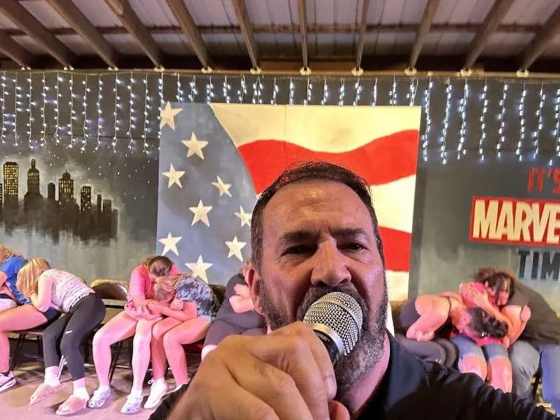 Hypnotist with microphone and American flag backdrop, participants appear hypnotized in a live show.